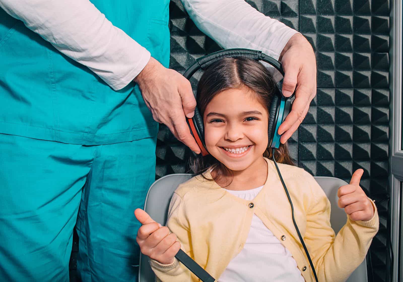 child smiling giving double thumbs up as audiologist fits head set for hearing test
